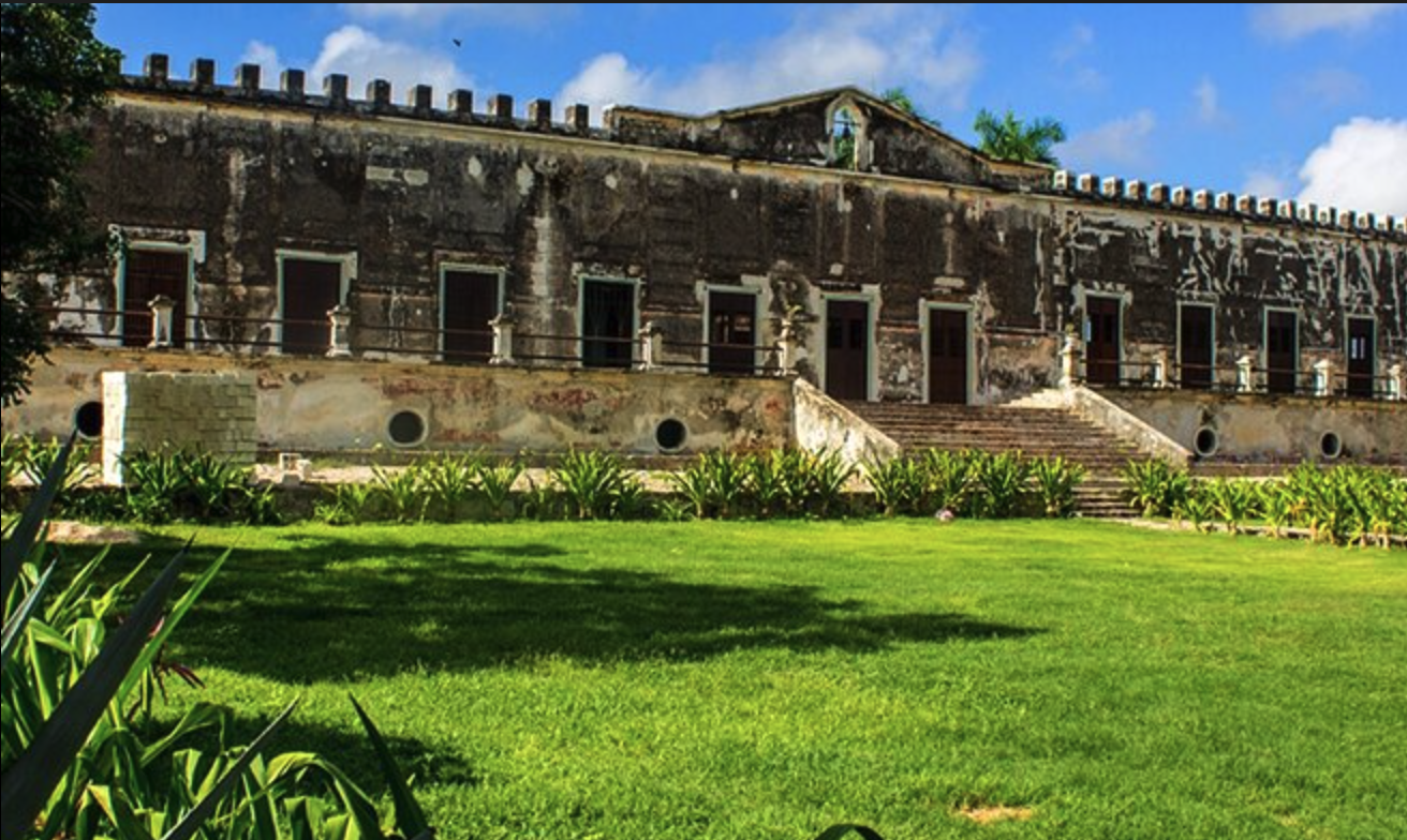 Historic hacienda facade with weathered stone walls, crenellated roofline, symmetrical dark wooden doors, and a stone staircase, situated behind a vibrant manicured lawn with low green shrubbery under a bright sky.