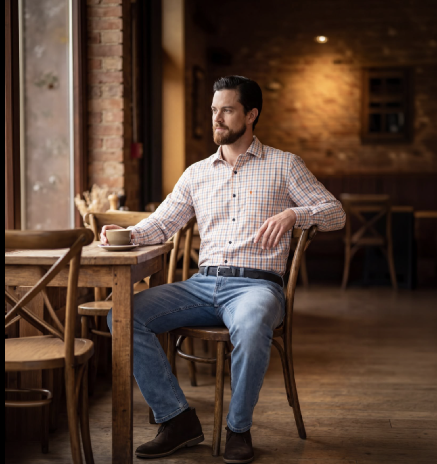 A warm, rustic interior of a cafe featuring exposed brick walls, dark wooden furniture, and a large window to the left providing natural light.